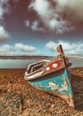 Weathered, small traditional Aiola boat in Seixal Bay, Portugal. Old wooden boat with blue and red paint chipped and faded. Sandy shore and a calm body of water and a cloudy sky, with a warm, vintage color palette. tranquility and nostalgia.