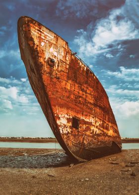 Large, old rusted shipwreck in Seixal Bay, Portugal. Ship run aground or beached, rusting on a sandy shore low-tide. Salvage of scrap metal to recycle on the shipyard recycling industry. Background features a bright blue sky with fluffy white clouds.