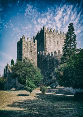 Guimaraes, Portugal. Medieval Guimaraes Castle, the most known castle in Portugal as it was the birthplace of the first Portuguese King. High towers and crenellated walls made of stone. The castle is surrounded by trees, including a tall cypress tree.